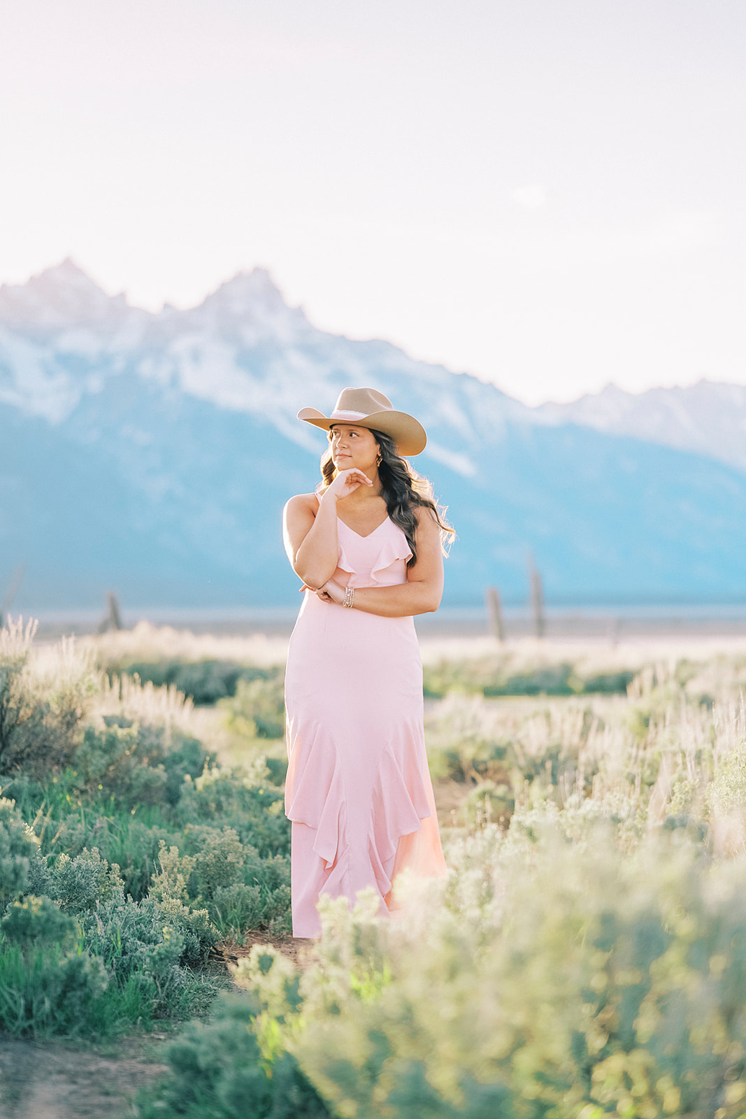 A woman in a pink dress and tan cowboy hat stands with hand under her chin while walking a trail near the mountains