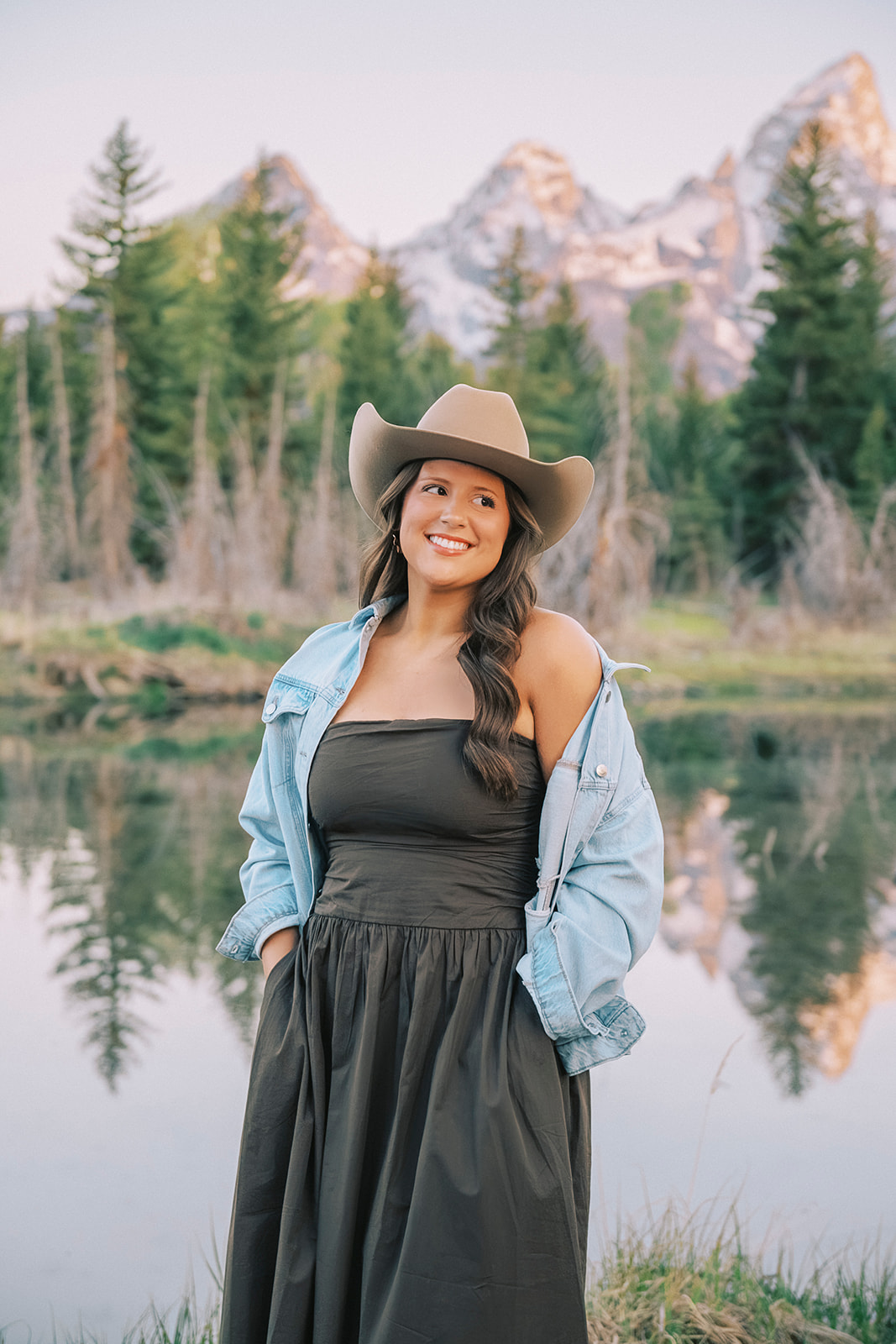 A happy high school senior in a dark dress and cowboy hat walks with hands in pockets by the river for her senior portraits in Jackson Hole