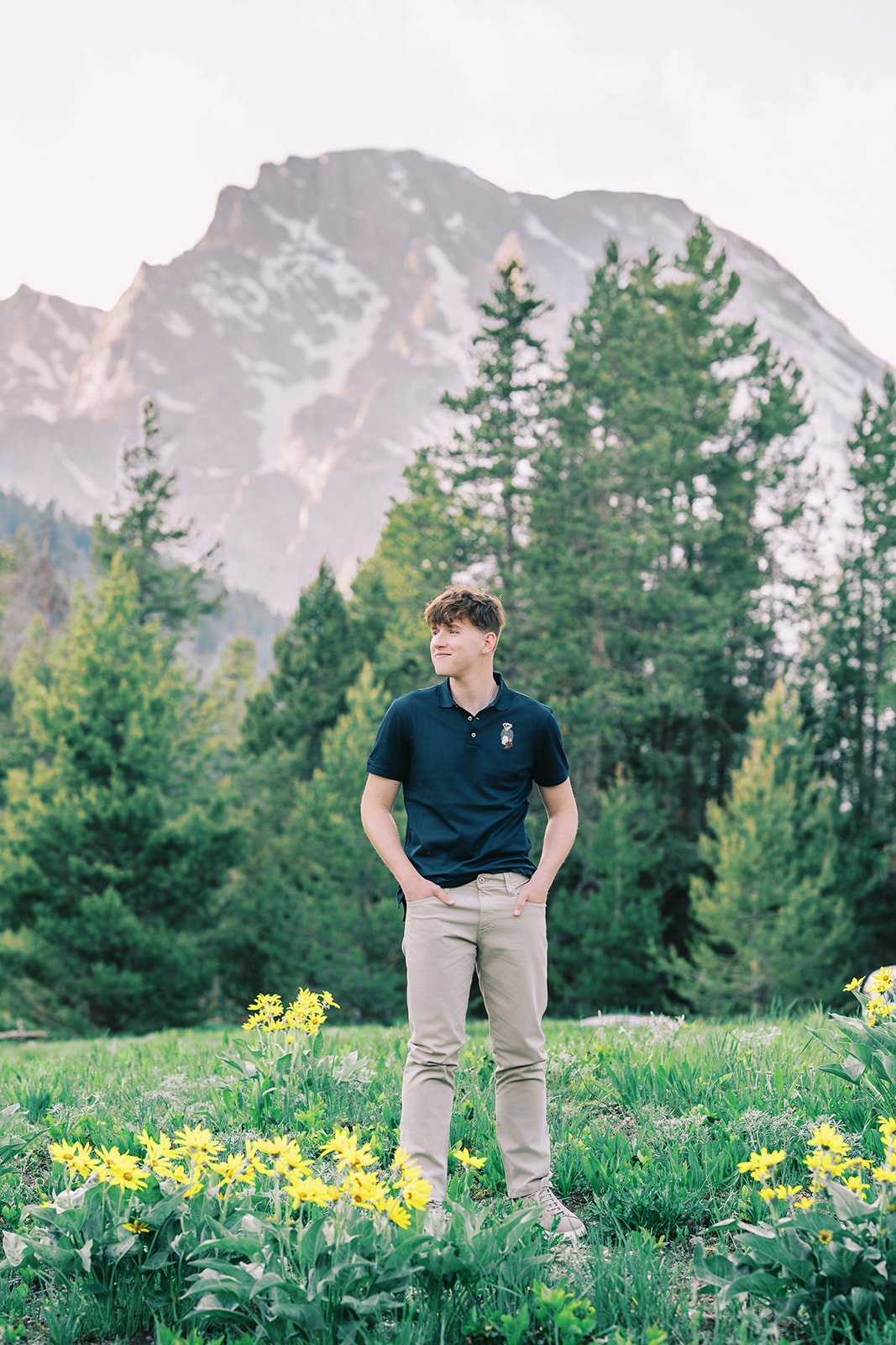 A man in a blue polo and khakis stands with hands in his pockets in a meadow with yellow wildflowers for his senior portraits in Jackson Hole