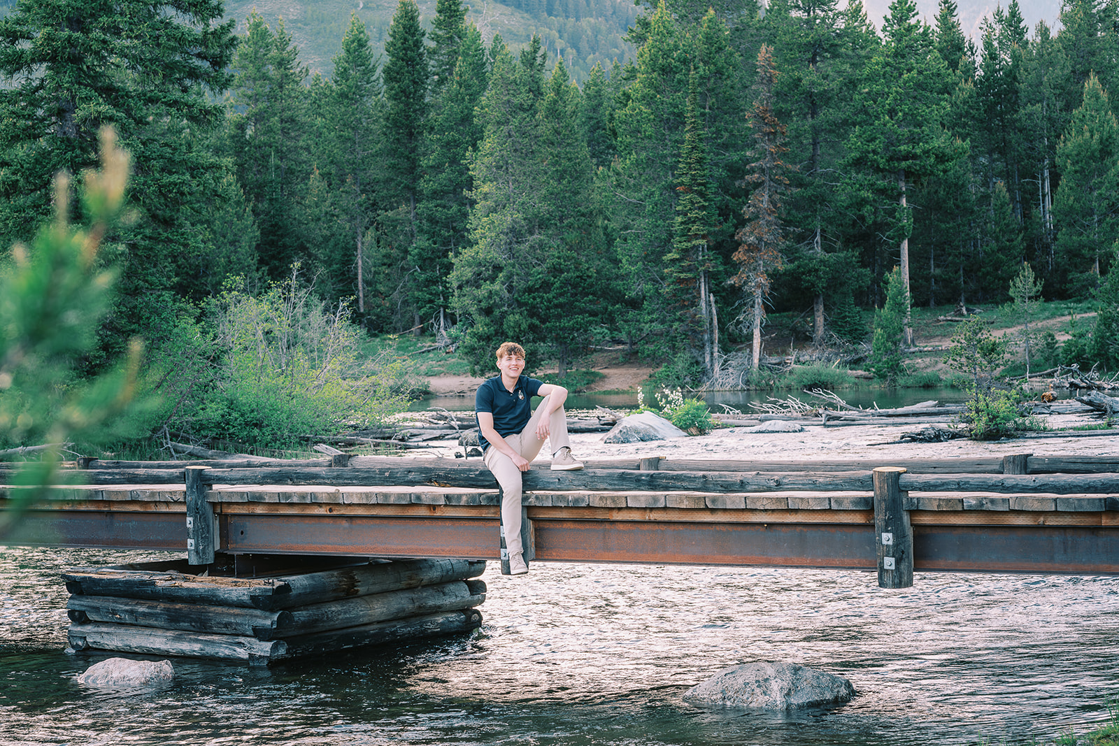 A laughing teenager in a blue polo and khakis sits with one leg dangling off the edge of an old bridge over a river