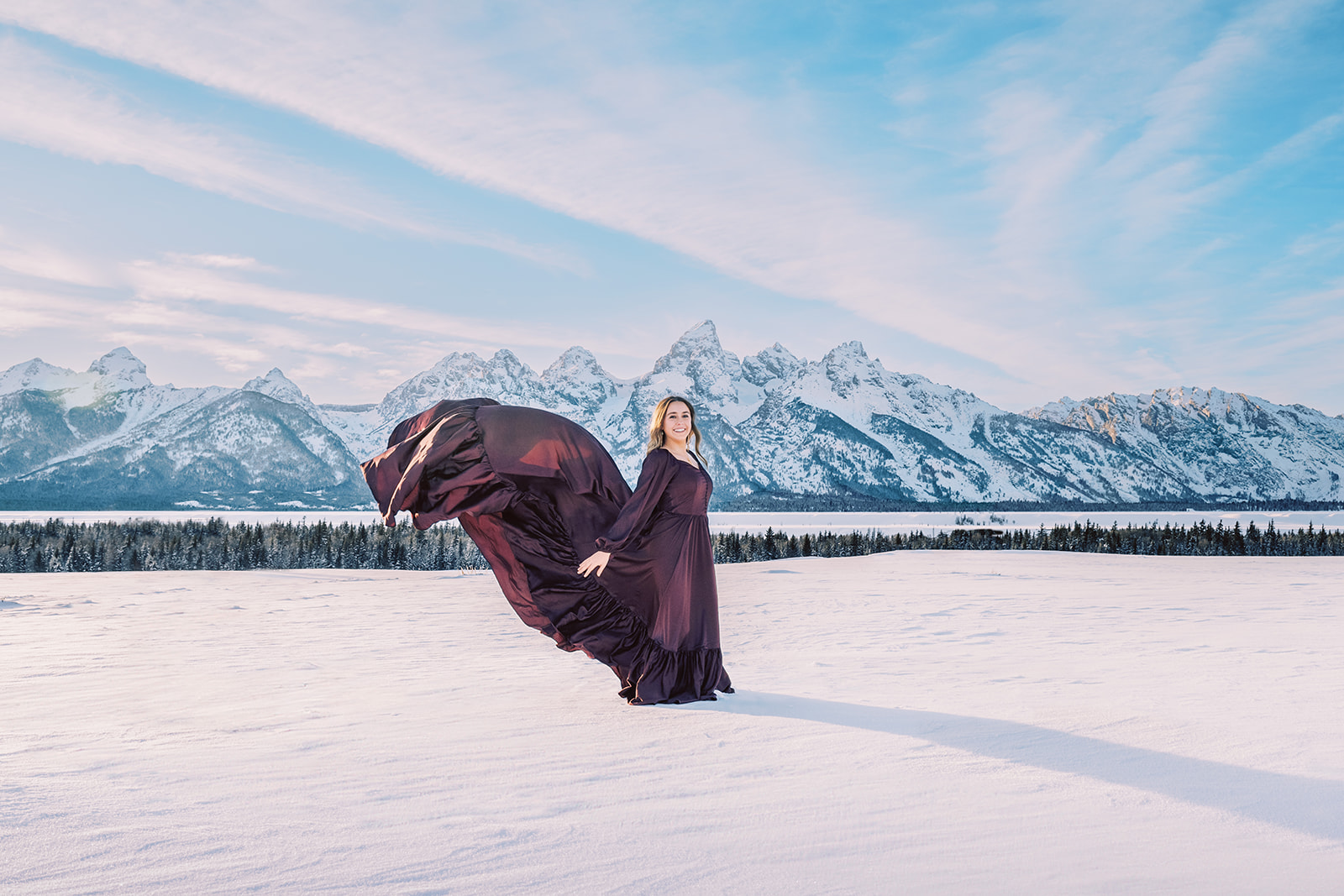A smiling woman stands in a snow covered field for senior portraits in Jackson Hole in a long purple dress with the train flowing in the wind behind her at sunset