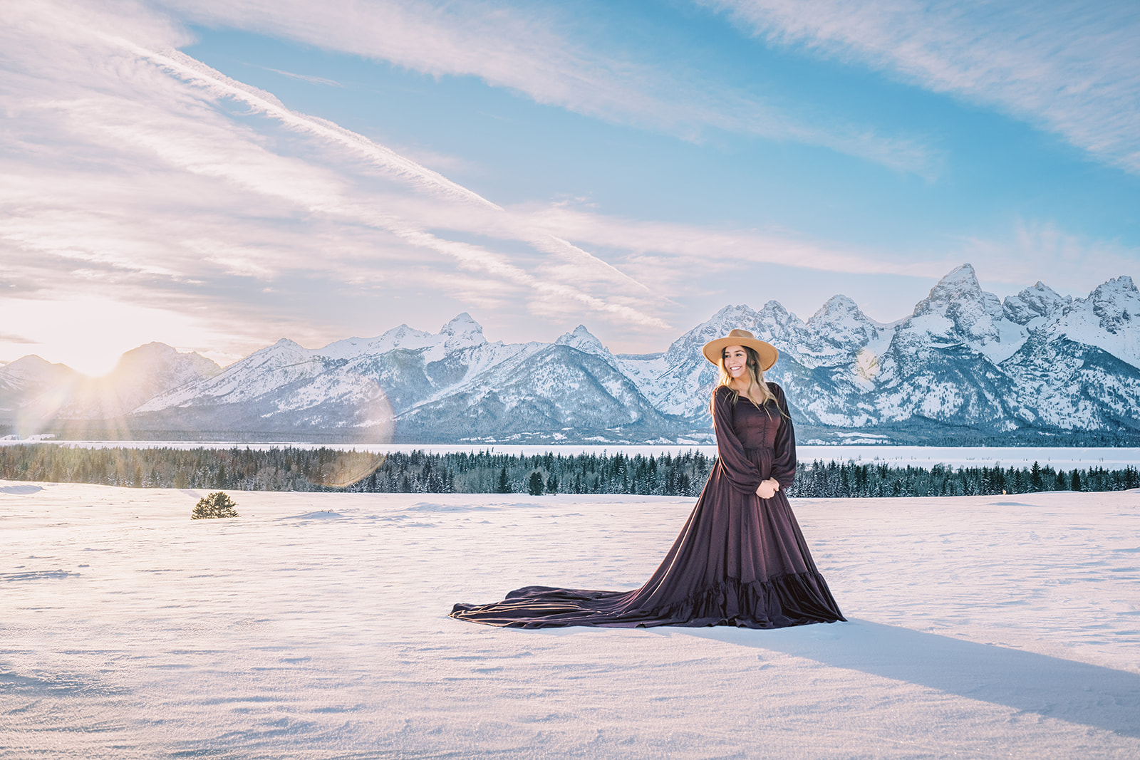 A high school senior in a long purple gown smiles while walking on snow near the mountains during her stunning senior portraits in Jackson Hole