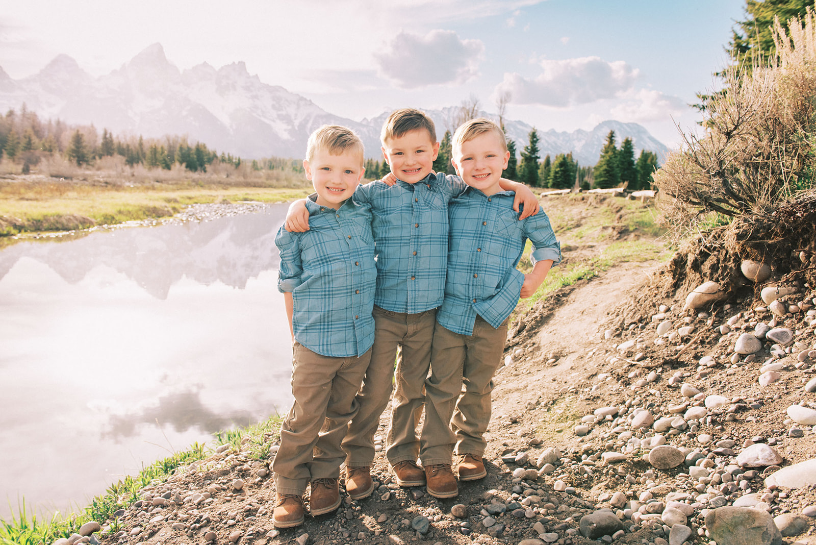 Three young brothers in matching blue shirts and khaki pants stand with arms around each other smiling on a lakeside trail during things to do in spring in Jackson Hole