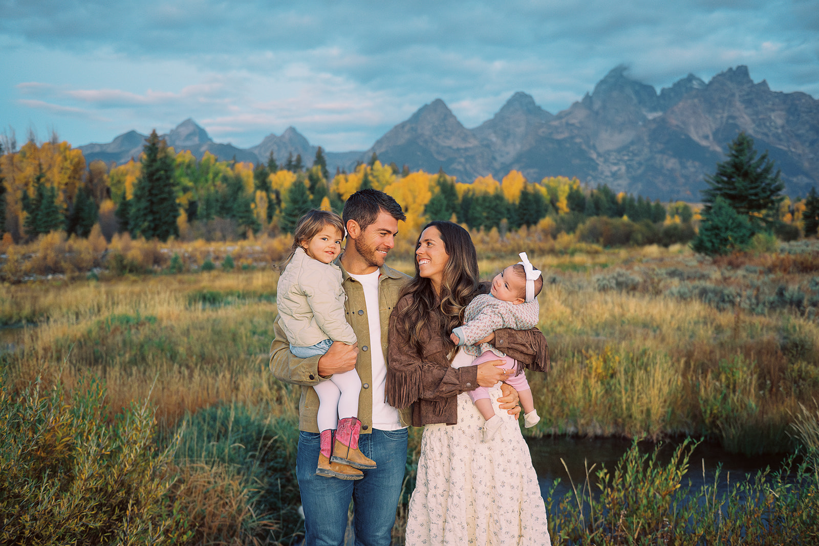 Smiling mom and dad look at each other while holding their toddler daughters by a river
