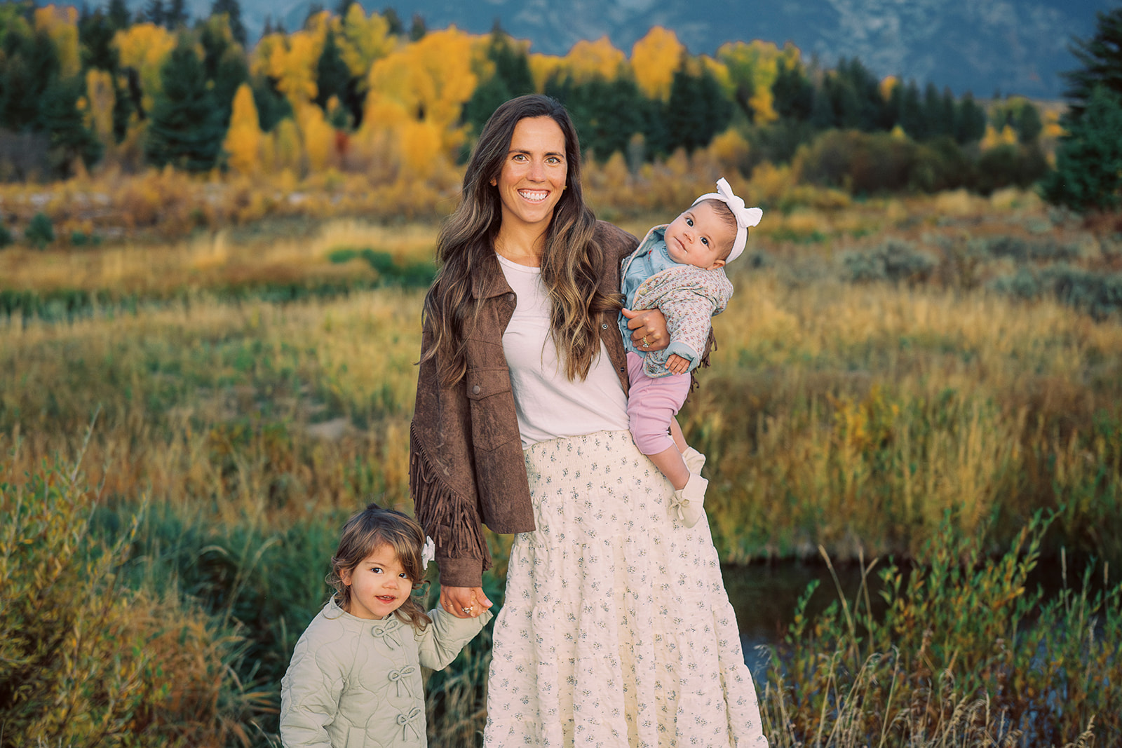 A smiling mother in a cream dress and brown jacket stands with her baby on her hip and toddler holding her hand in a trail at sunset