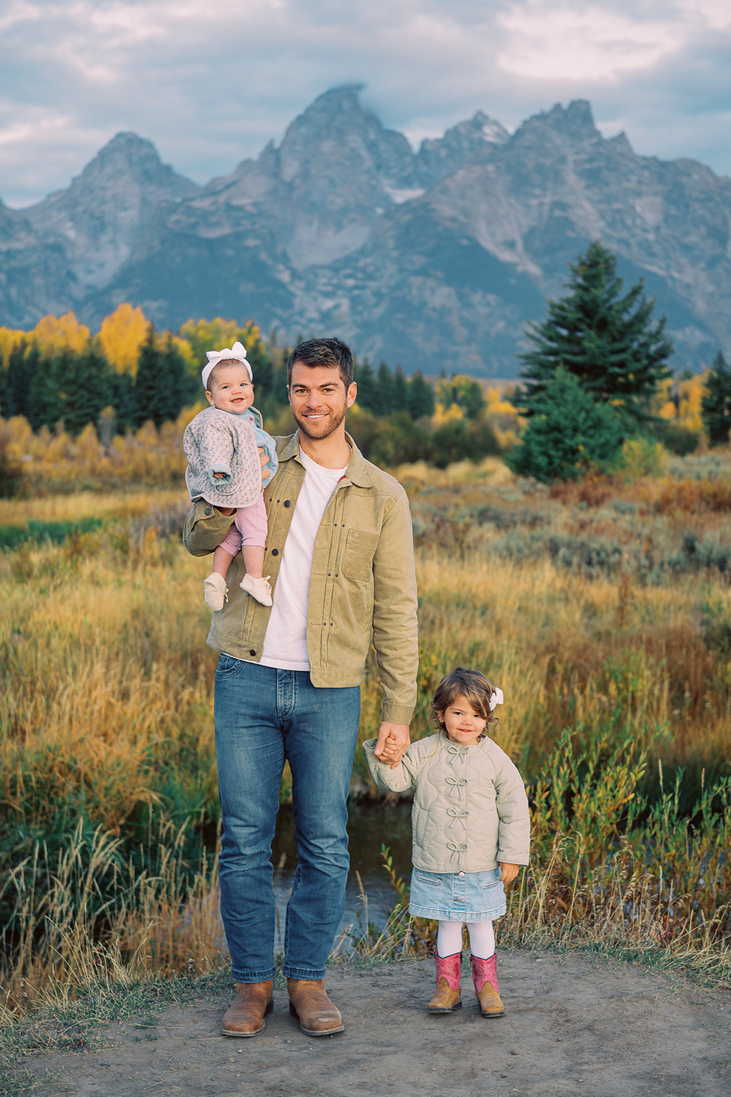 A happy dad in jean and tan jacket stands holding hands with his matching two toddler daughters in a meadow after finding things to do in in Grand Teton National Park in Fall
