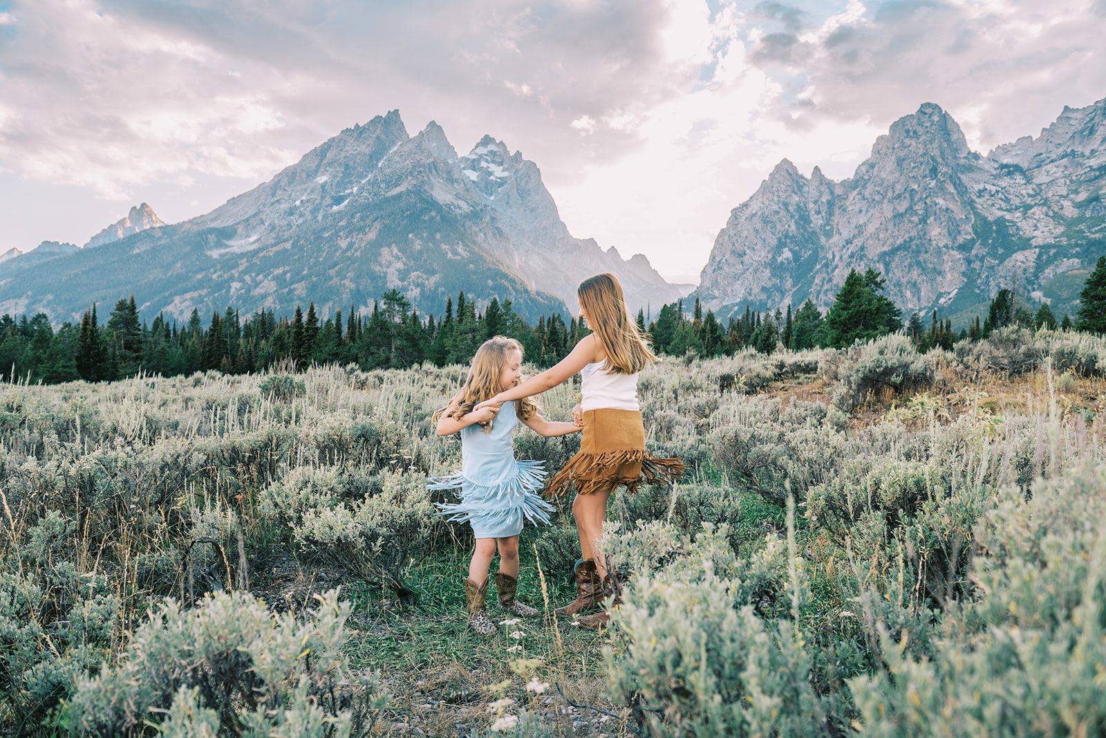 Happy young sisters in fringe dresses dance and twirl while holding hands in a mountain meadow at sunset