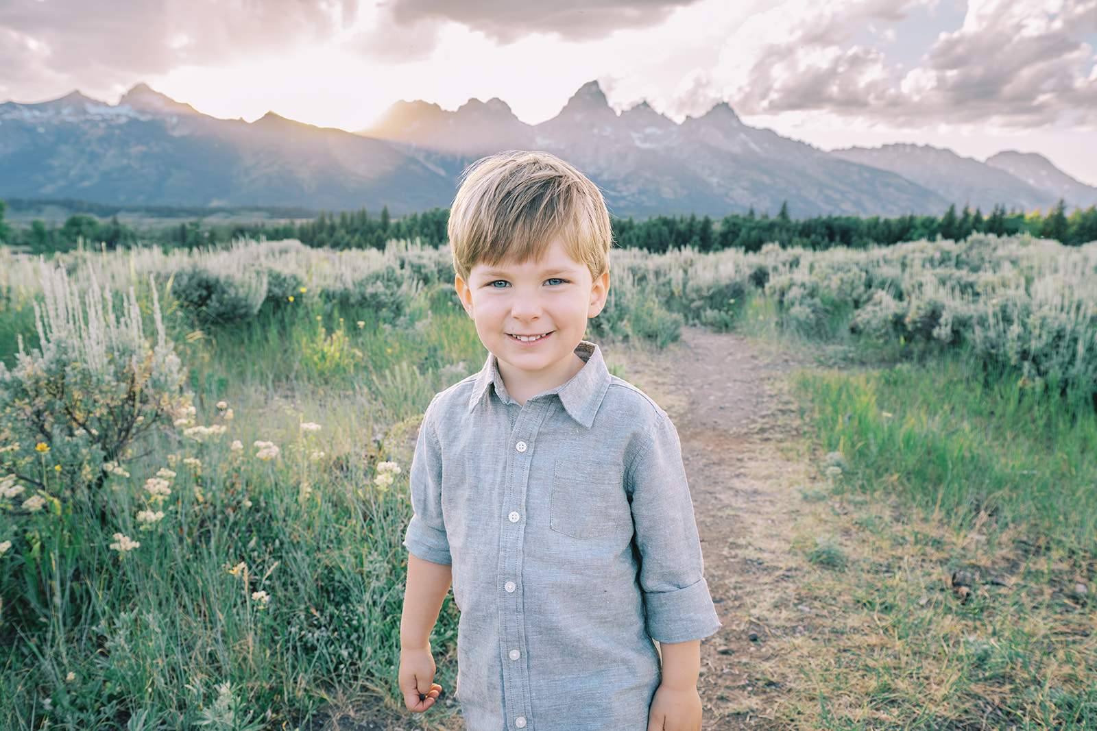 A closeup photo of a smiling toddler boy in a grey button down walking in a meadow trail at sunset