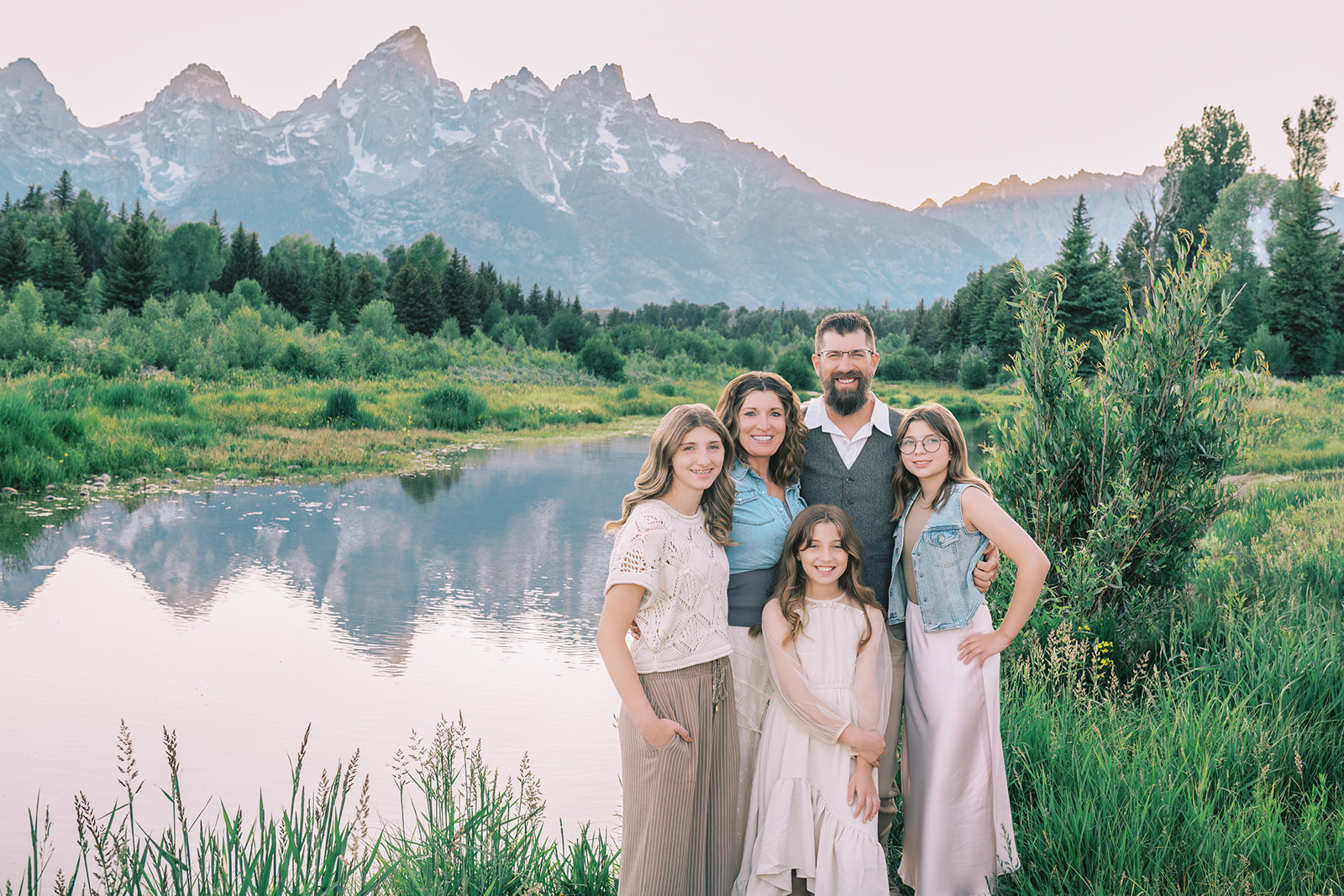 A happy family of five with three young daughters stands smiling by a river at sunset in the mountains