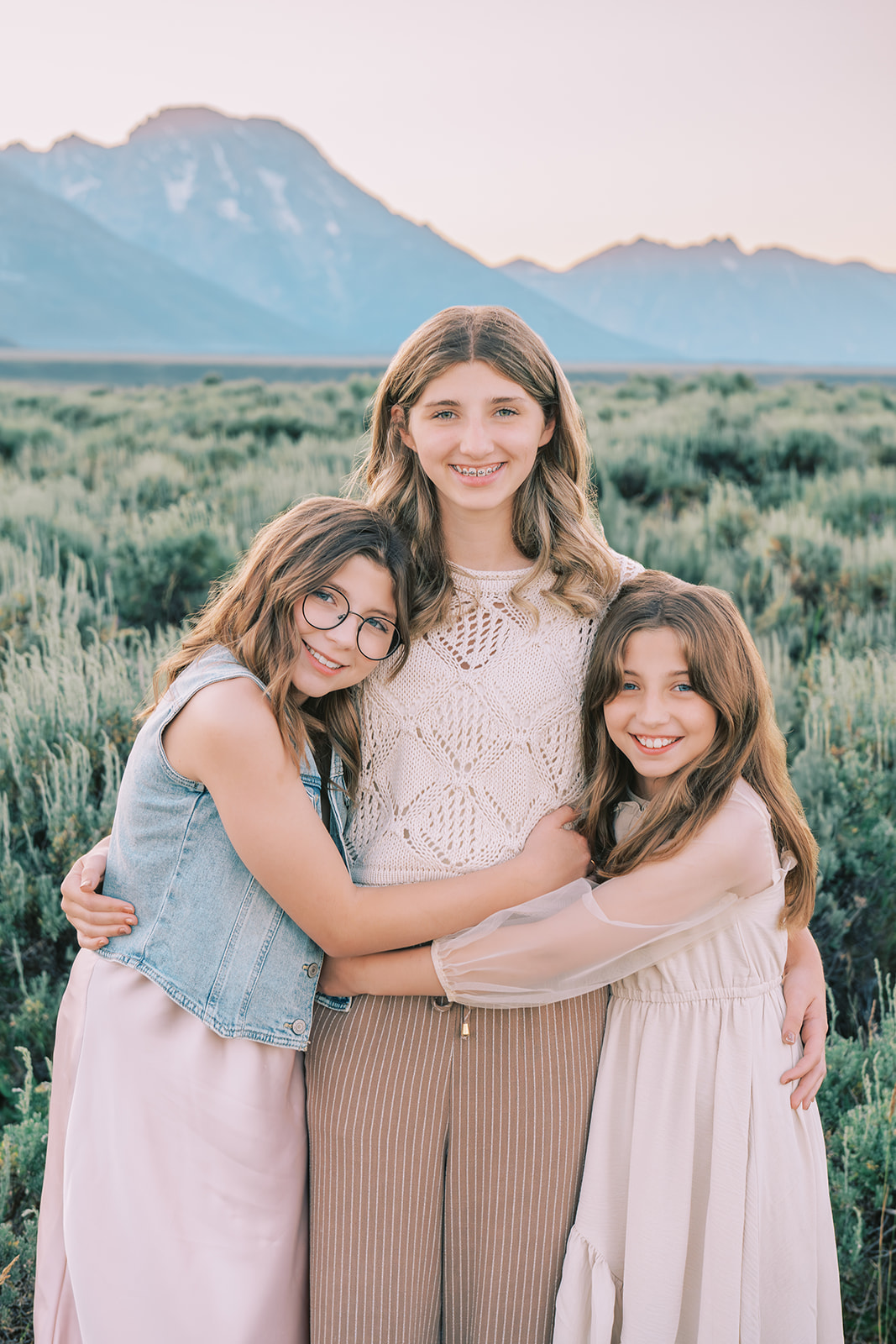 A teen girl stands smiling hugging her two younger sisters in pink dresses in a meadow at sunset