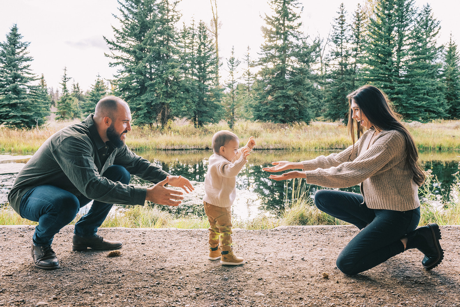 Happy mom and dad reach for toddler to walk to them in opposite directions in a trail by a river