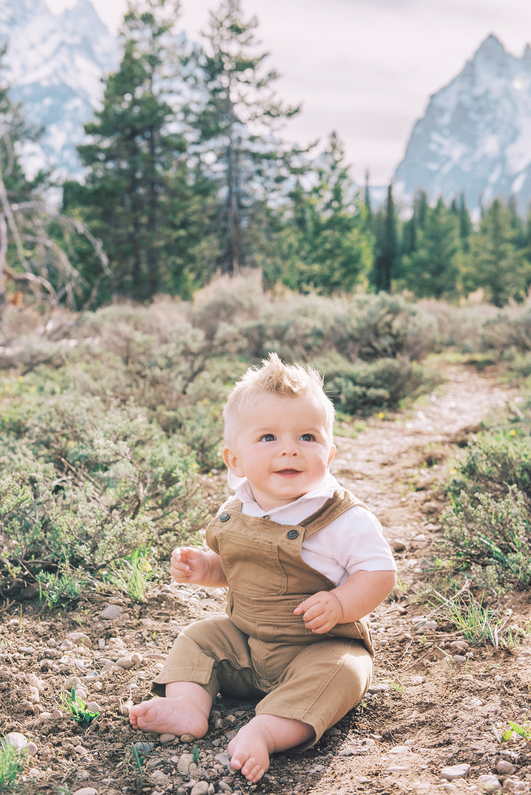 A toddler boy in tan overalls and white shirt sits barefoot in a mountain trail while smiling after mom learned what to wear for family photos