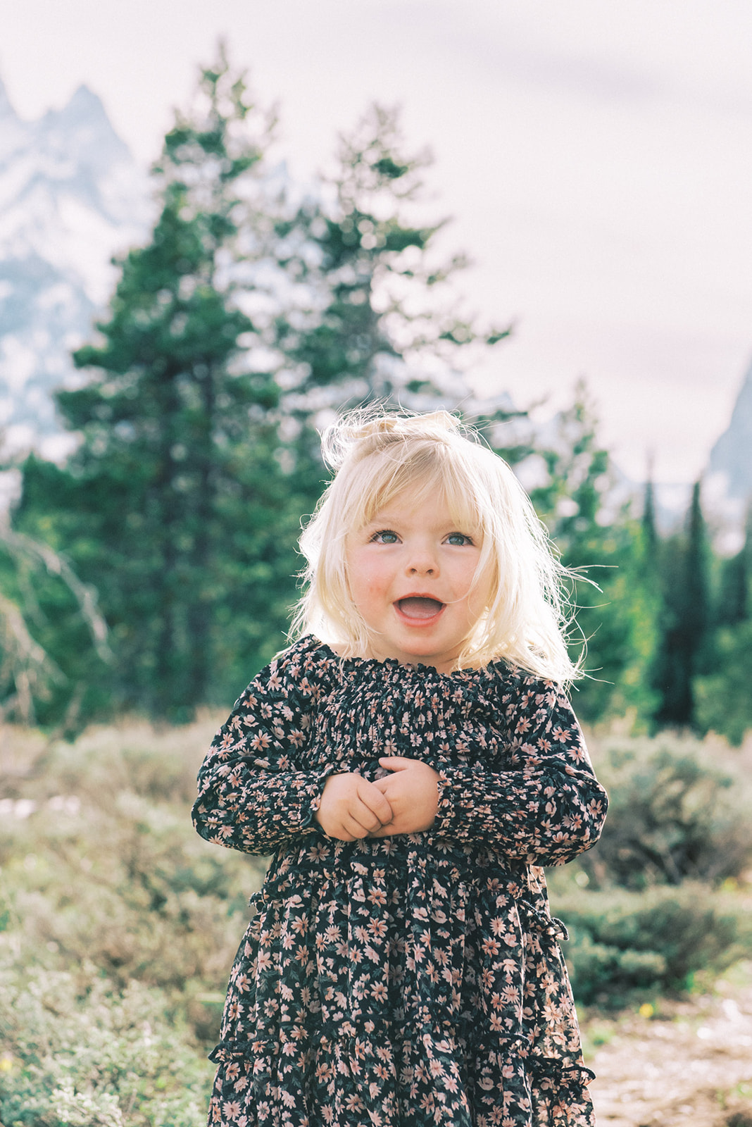 A young girl in a flower print dress smiles big while exploring a mountain trail after mom learned what to wear for family photos