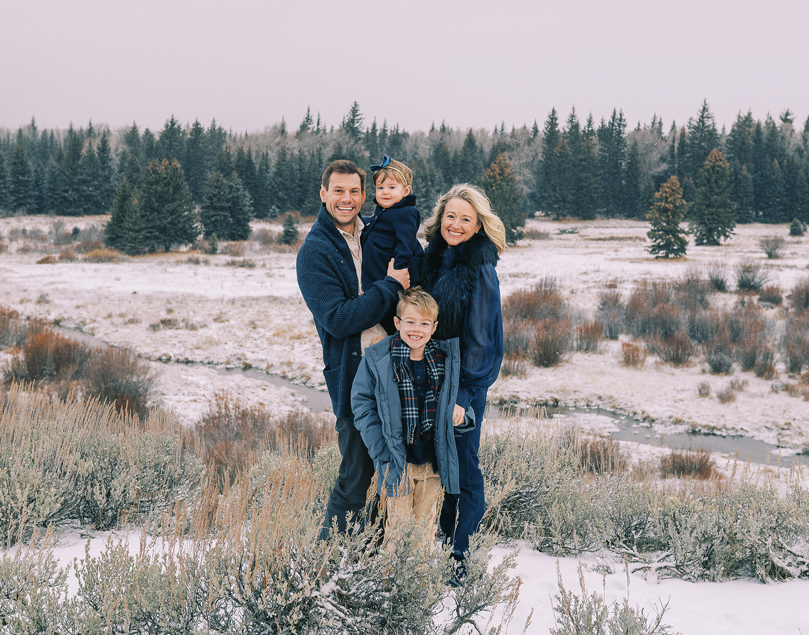 A happy family with a toddler girl in dad's arms and young boy at their feet stand in a snow covered meadow after learning what to wear for family photos