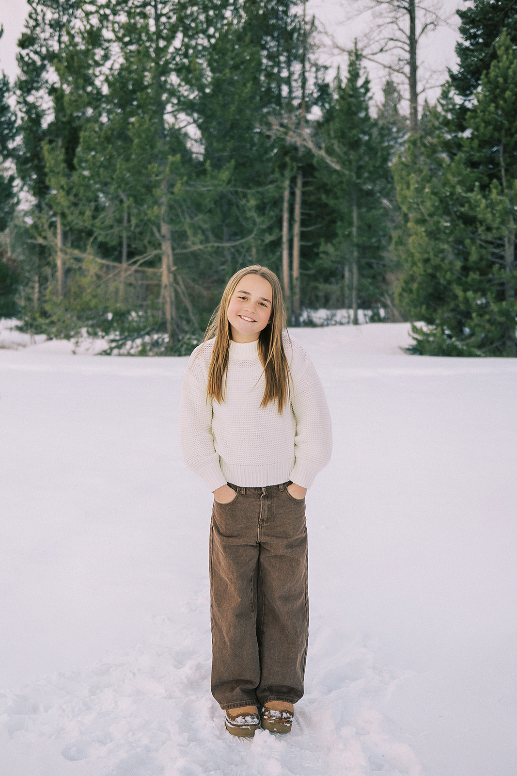 A young girl in brown pants and white sweater stands with hands in pockets while smiling in a snow covered forest trail