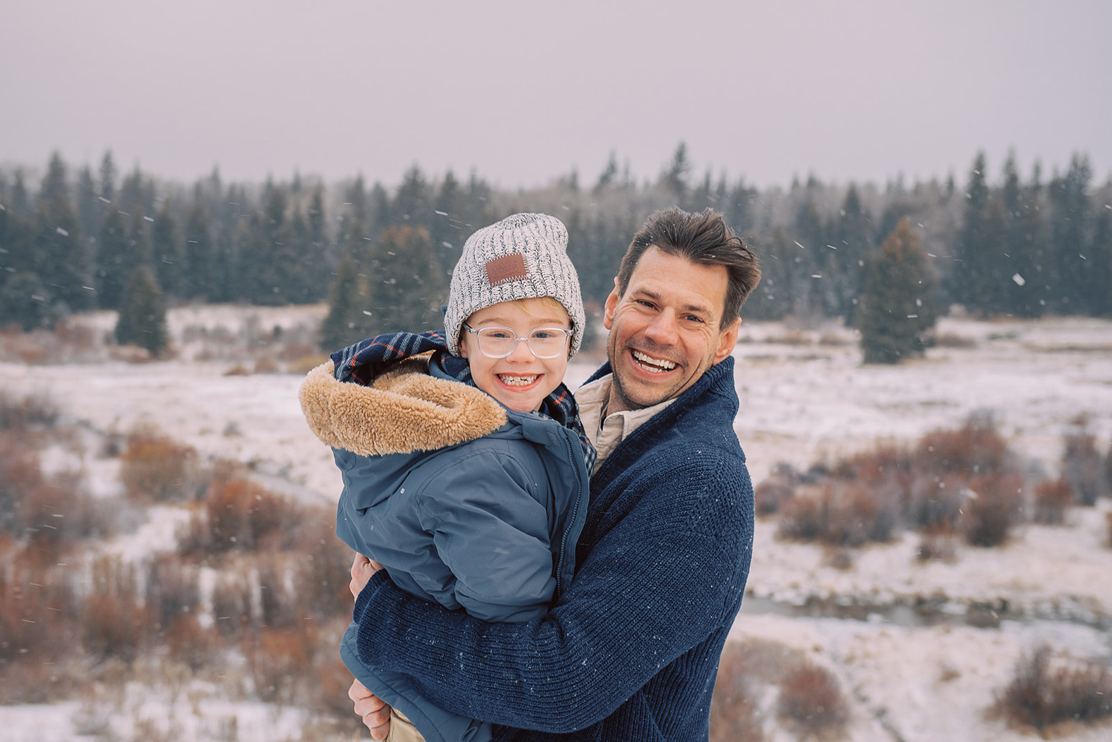 A smiling father holds his young son in a snow covered meadow wearing thick blue coats for their winter family photo outfits
