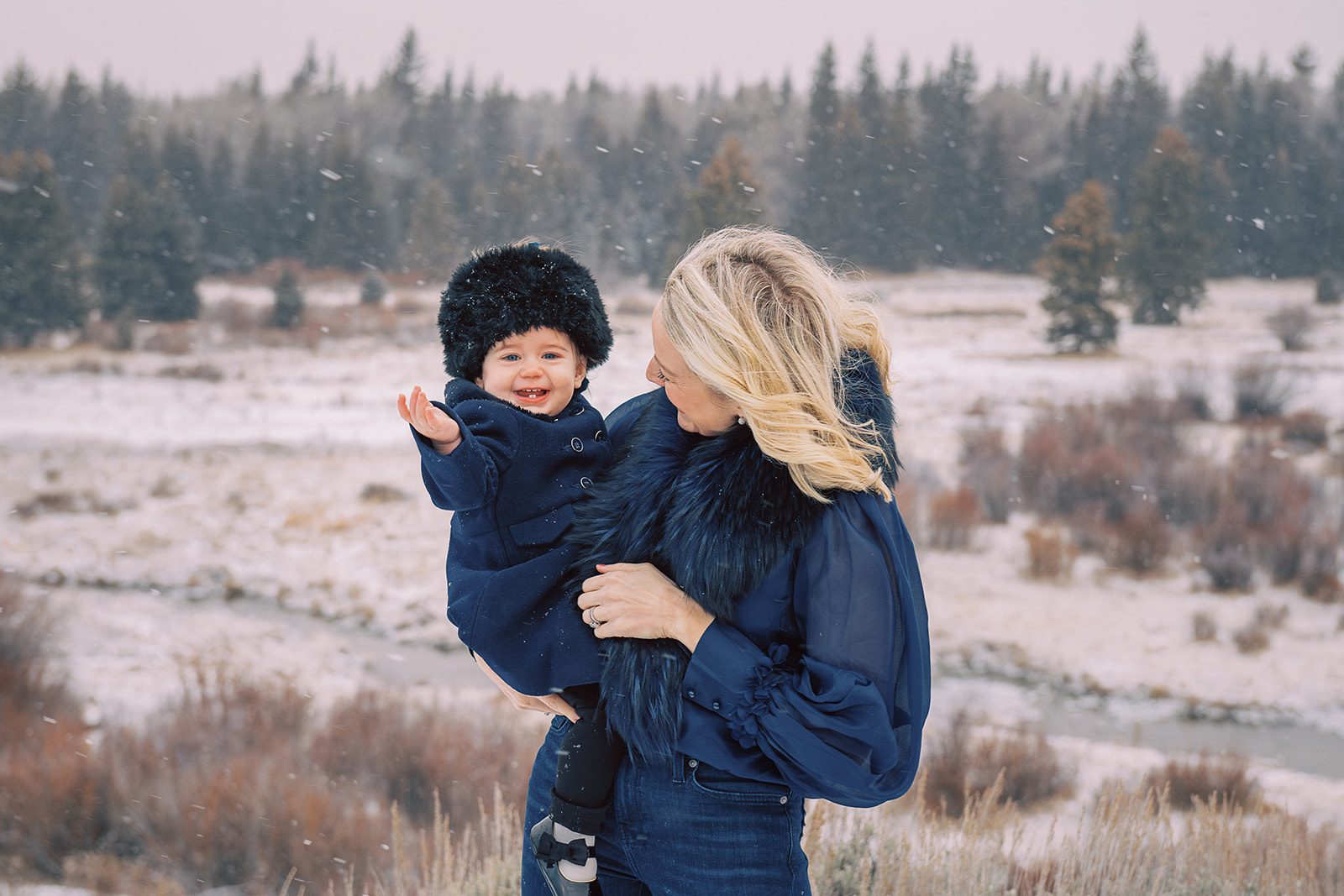 A smiling toddler girl in a blue coat and furry cap sits on mom's hip in a snowy meadow after shopping for winter family photo outfits