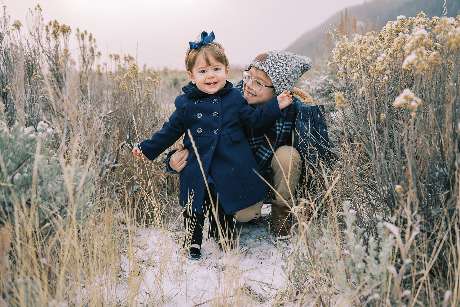 A toddler girl and her big brother explore snow covered trail through grasses in blue winter family photo outfits