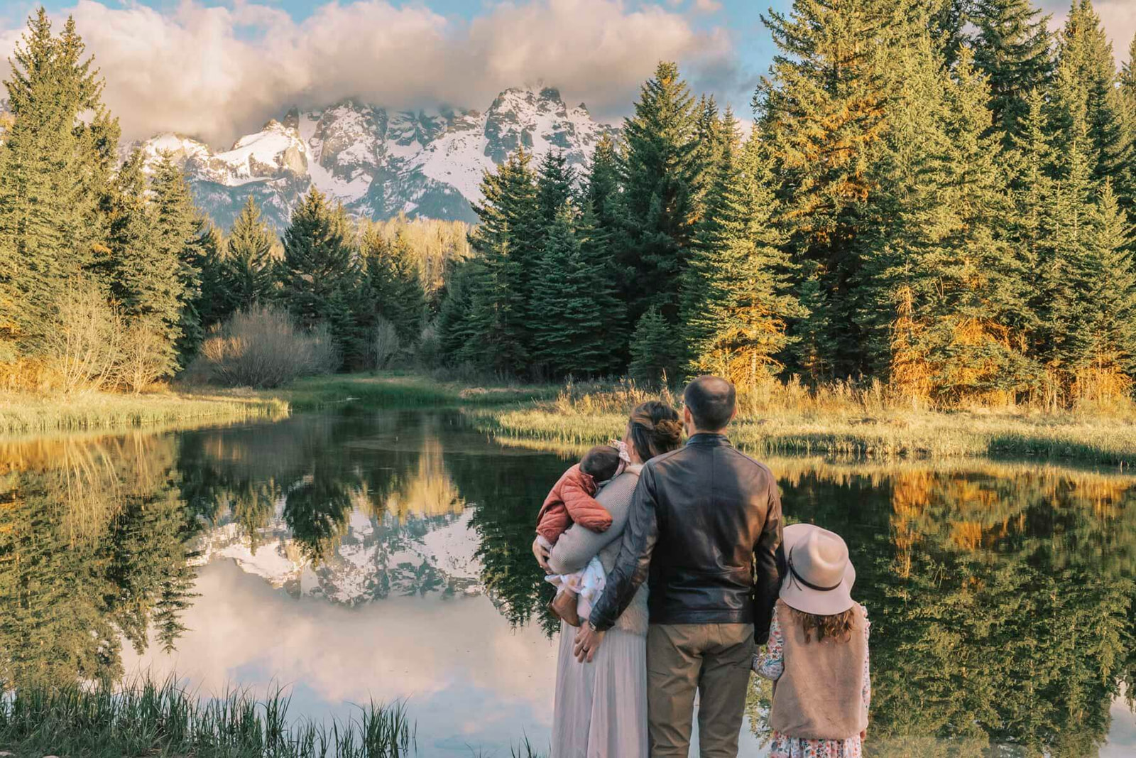A mom and dad admire snow covered mountains with their baby and toddler daughters on the edge of a river