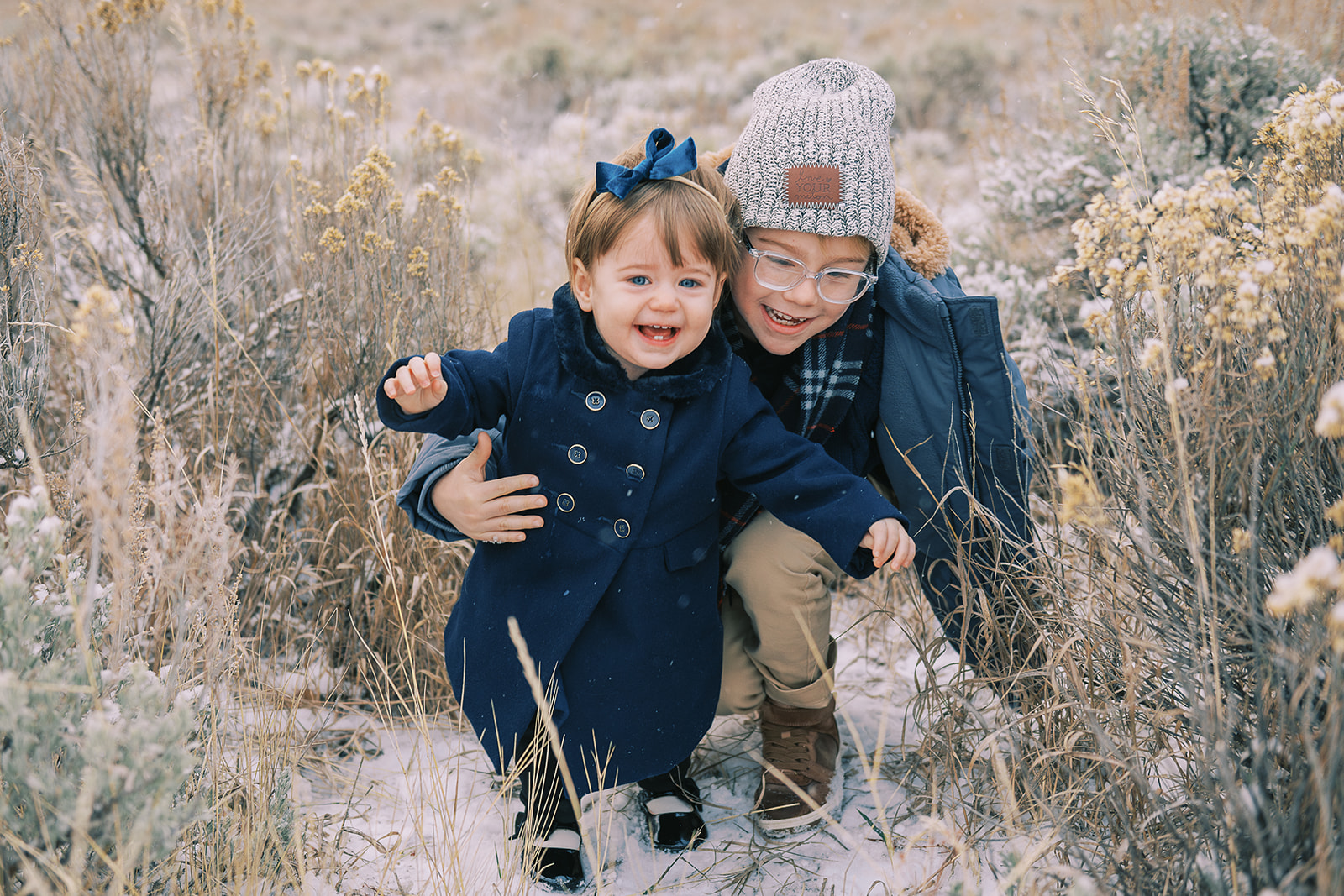 A toddler girl laughs with big brother who is helping her on a hike through snow covered pasture in blue coats during the best time to visit Jackson Hole