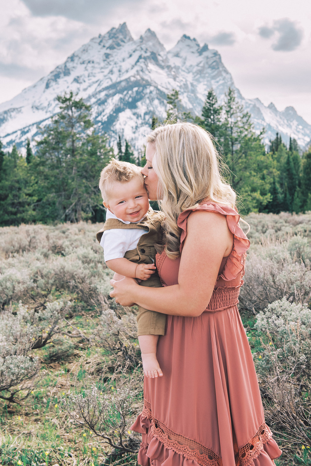 A happy mom in a pink dress kisses her toddler in tan overalls in her arms while on a hike and enjoying the best time to visit Jackson Hole