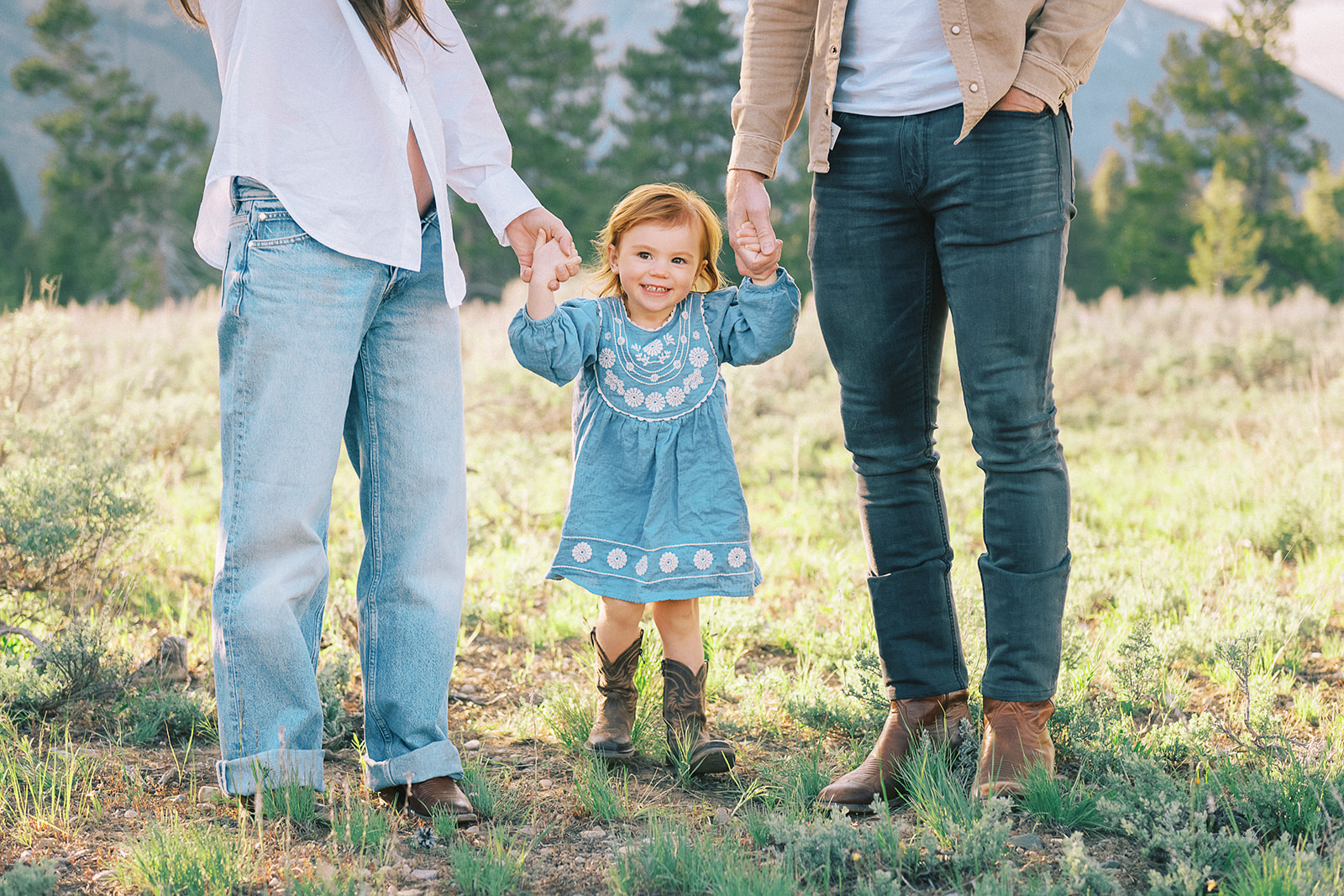 A giggling toddler girl in a blue dress walks with mom and dad holding their hands on a hill