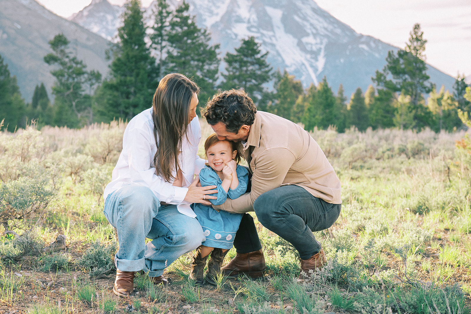 A toddler girl in a blue dress giggles while being tickled by mom and dad in a meadow at sunset after visiting children's boutiques in Jackson Hole