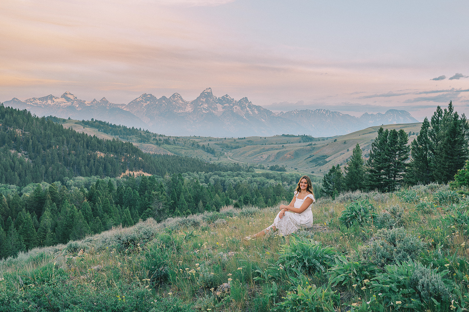 A happy woman in a white dress sits on a boulder on a hillside at sunset