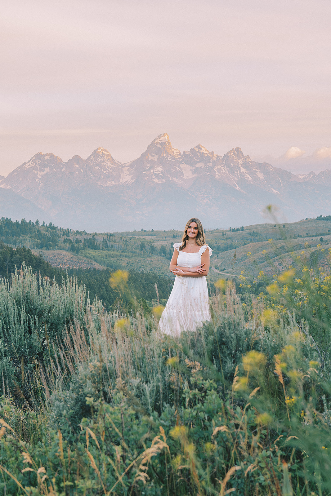 A high school grad stands on a hill overlooking a lush valley in a white dress with arms crossed