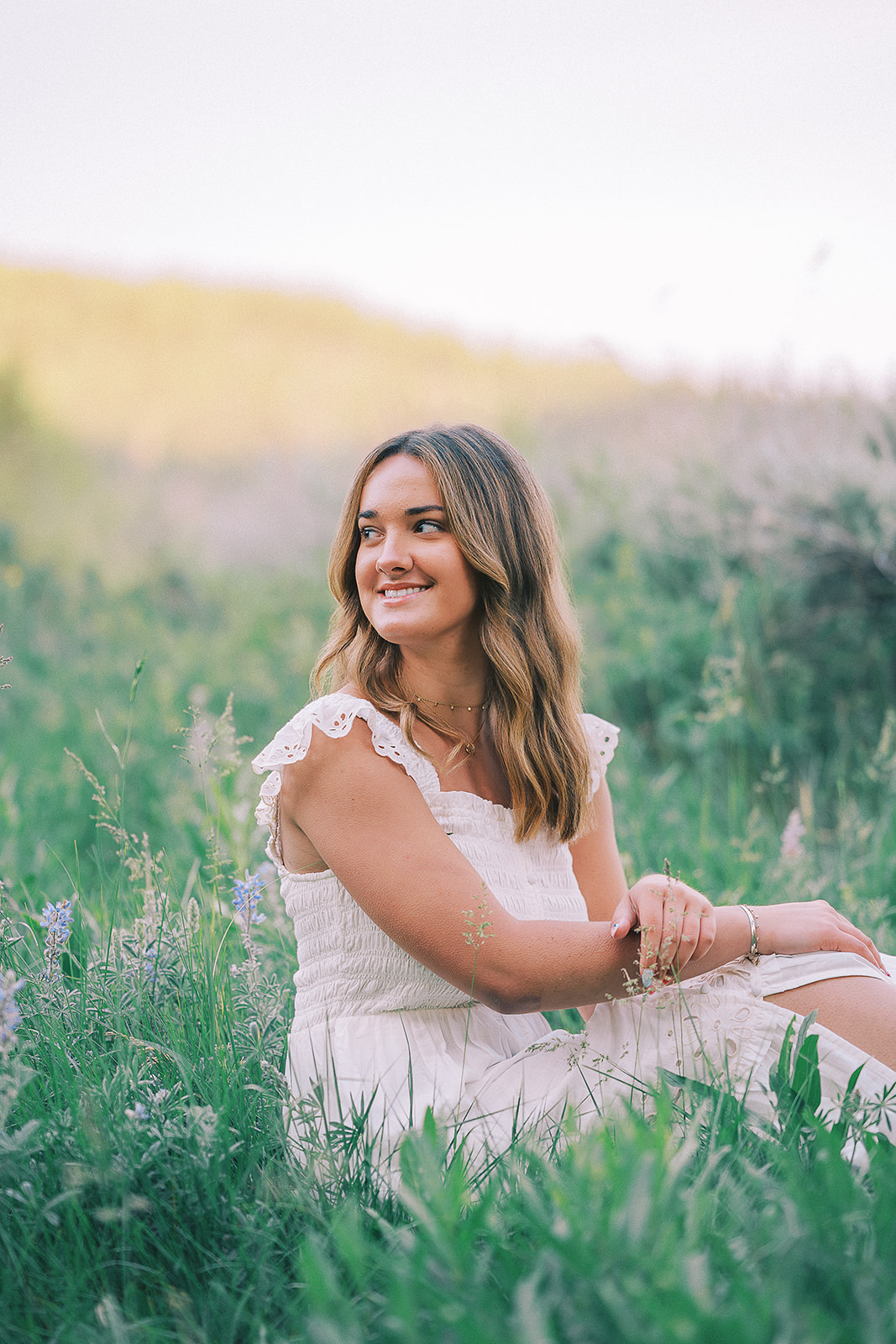 A happy woman in a white dress sits in wildflowers of a meadow at sunset smiling over her shoulder after researching college scholarships in Jackson Hole