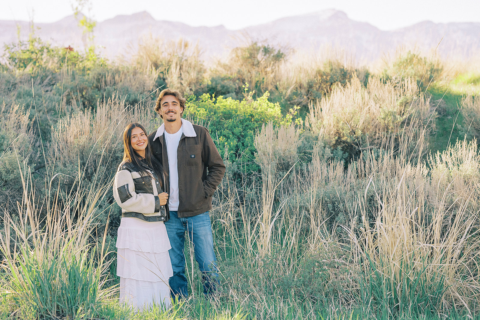 A couple in brown coats and white stand smiling together on a hike at sunset