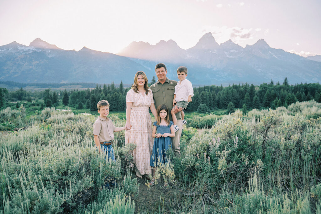 A happy family of 5 stand smiling in a meadow at sunset for some family portraits