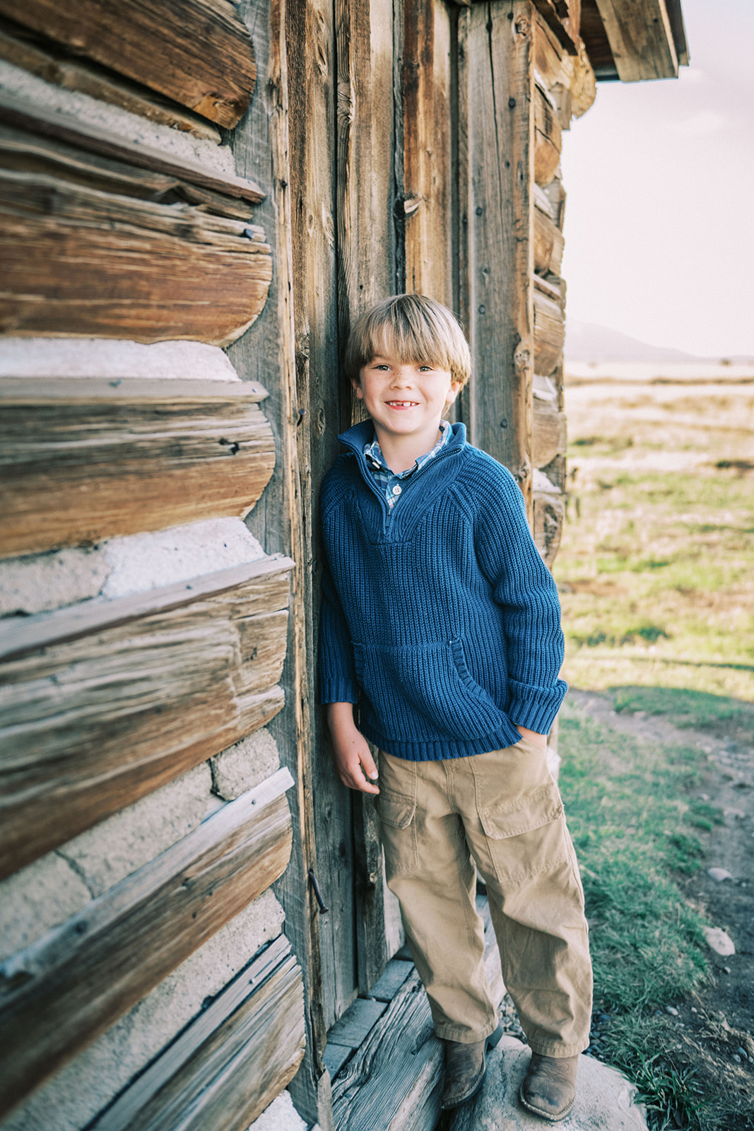 A young boy in a blue sweater smiles while leaning against a rustic old barn door