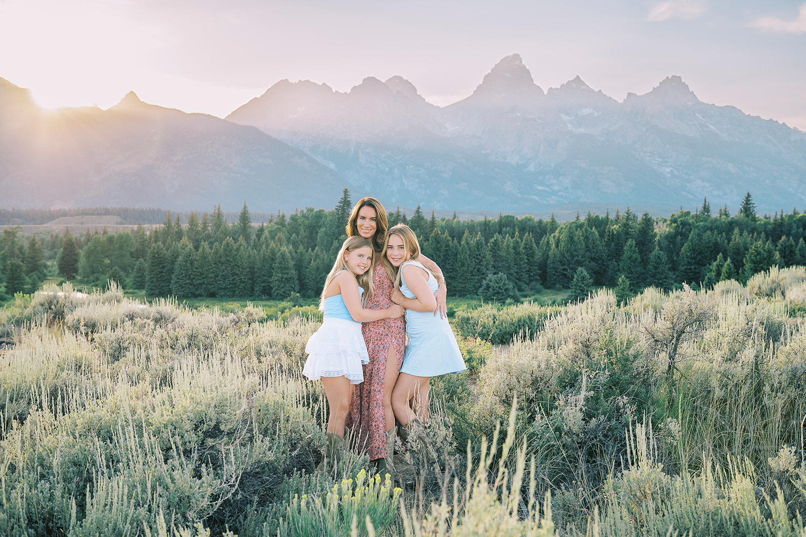 Happy mom in red floral print dress hugging her two young daughters in white and blue at sunset