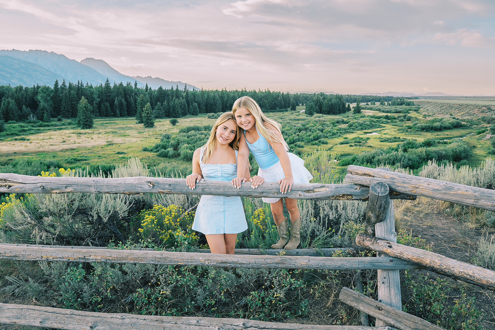 Two young blonde sisters in blue and white dresses climb a rustic old pasture fence overlooking a meadow after enjoying hair salons in Jackson Hole