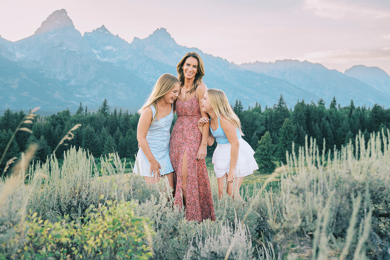 A laughing mom in a red dress stands with her giggling daughters in matching blue outfits in a forest after finding great hair salons in Jackson Hole