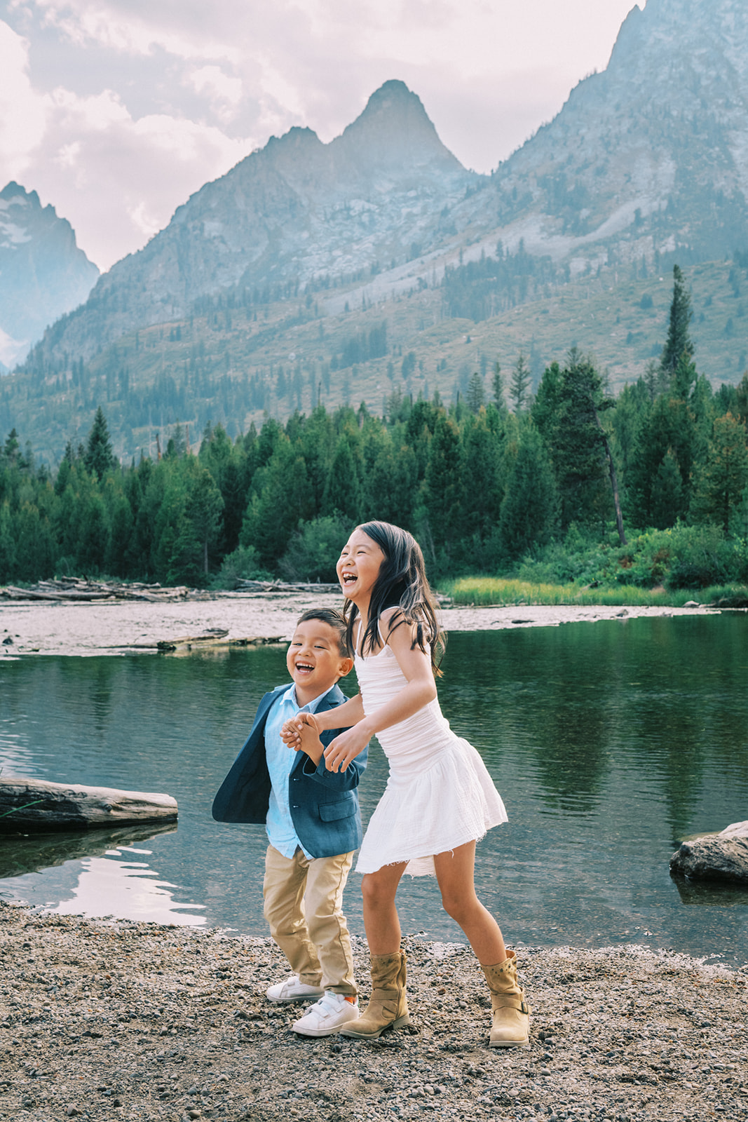 A happy brother and sister in white and blu laugh and play while holding hands on the edge of a river