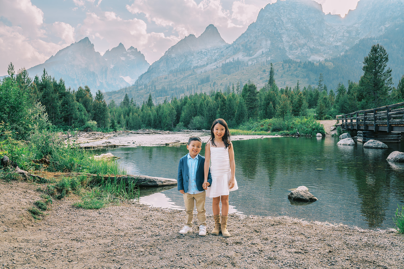 A happy young girl in a white dress stands holding the hand of her little brother on the edge of a river while staying at luxury lodging in Jackson Hole