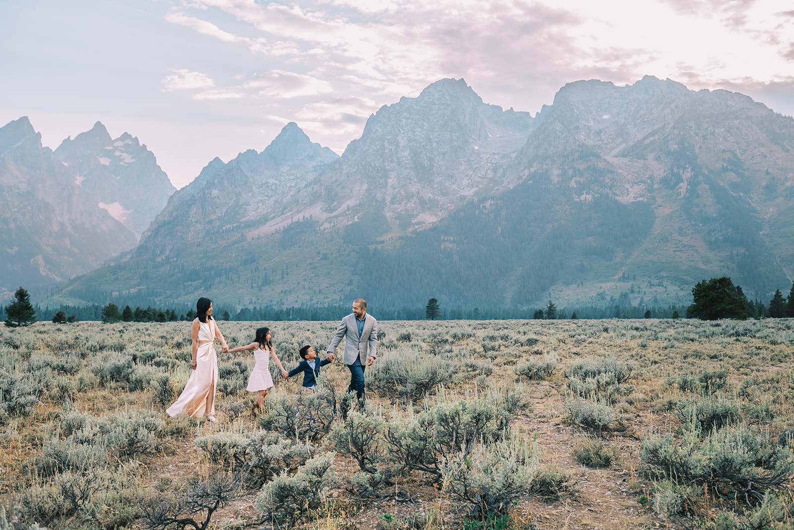 A dad in a grey suit jacket leads his wife and two toddlers by the hand in pink and blue on a hike after enjoying luxury lodging in Jackson Hole