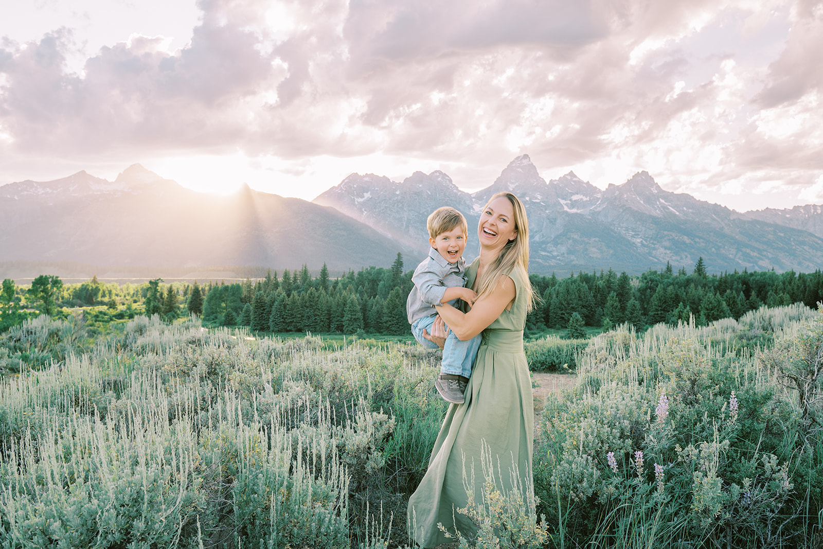 A laughing toddler sits in mom's arms in a meadow at sunset while she wears a green dress