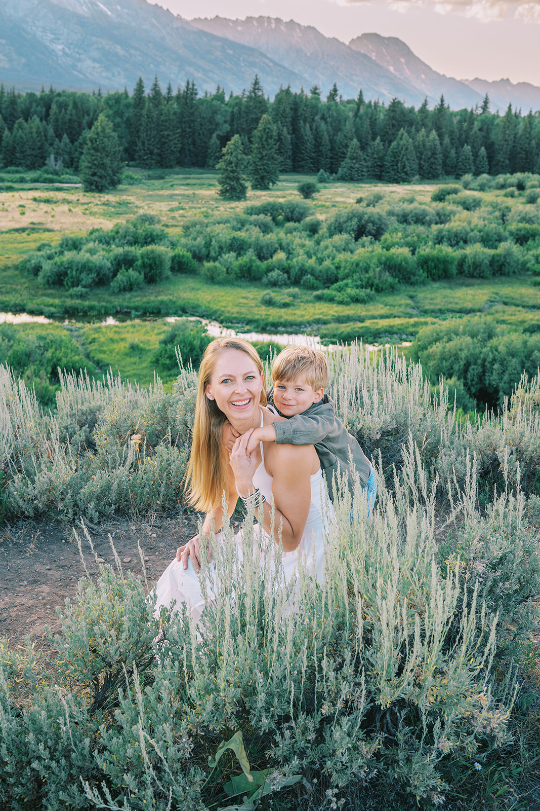 A young boy in a green jacket climbs on mom's back as she kneels in a white dress in a meadow after enjoying spas in Jackson Hole