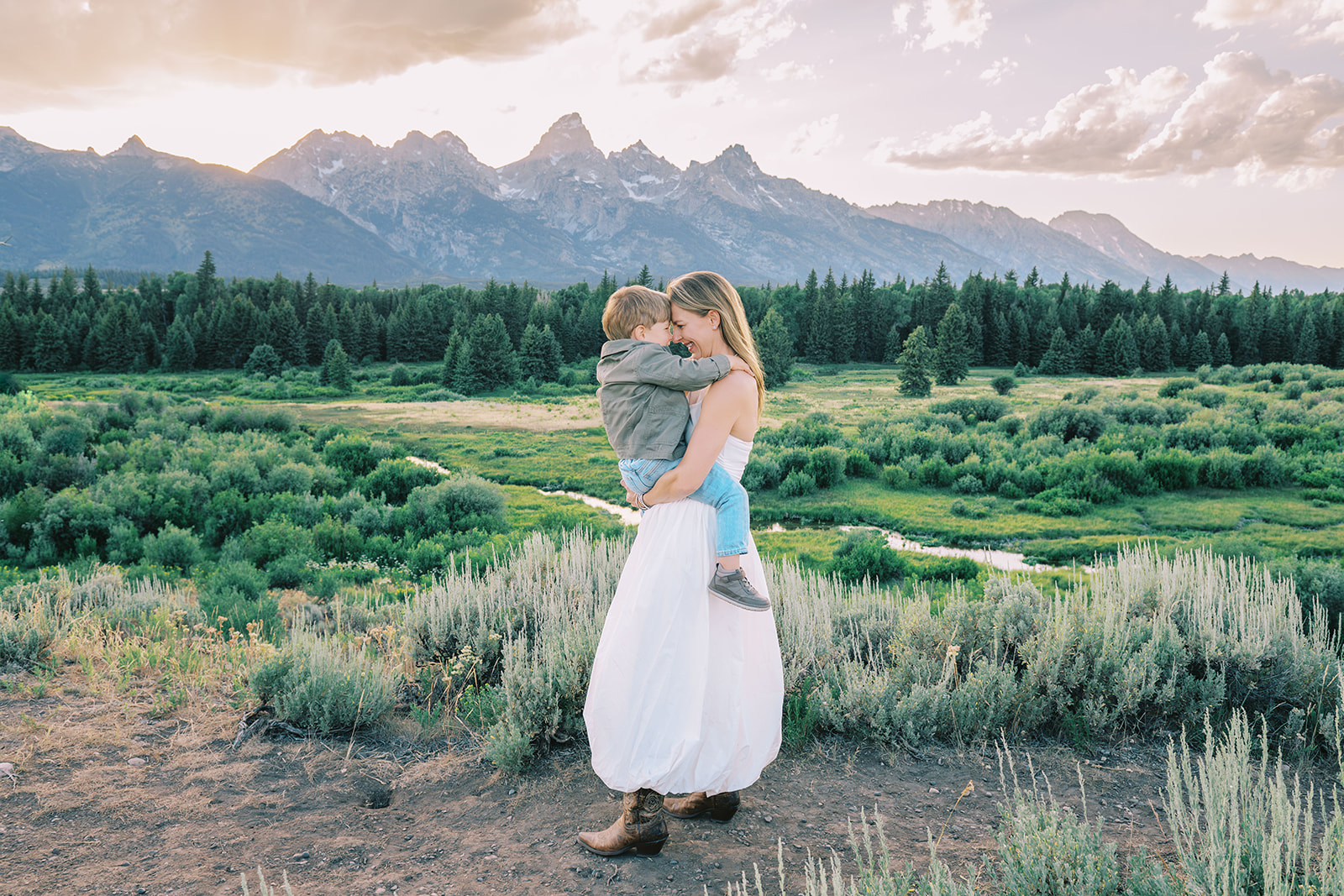 A happy mother in a white dress plays with her toddler son forehead to forehead in a trail at sunset after finding spas in Jackson Hole