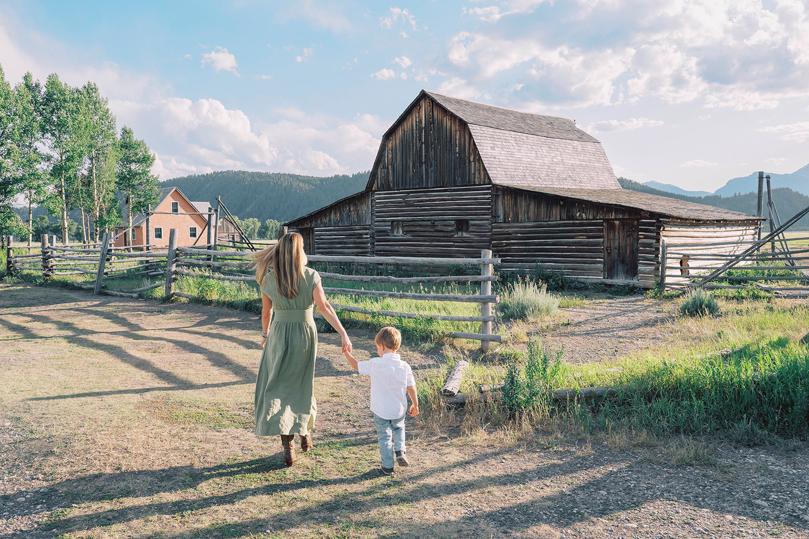 A mother in a green dress walks her young son in a white shirt on a path near an old barn after exploring great spas in Jackson Hole