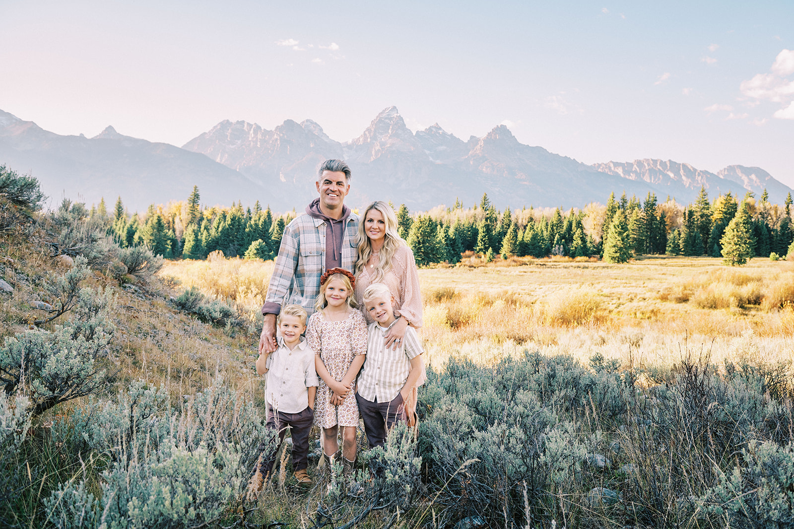 A happy mom and dad stand in a meadow at sunset with their three happy young children at their feet