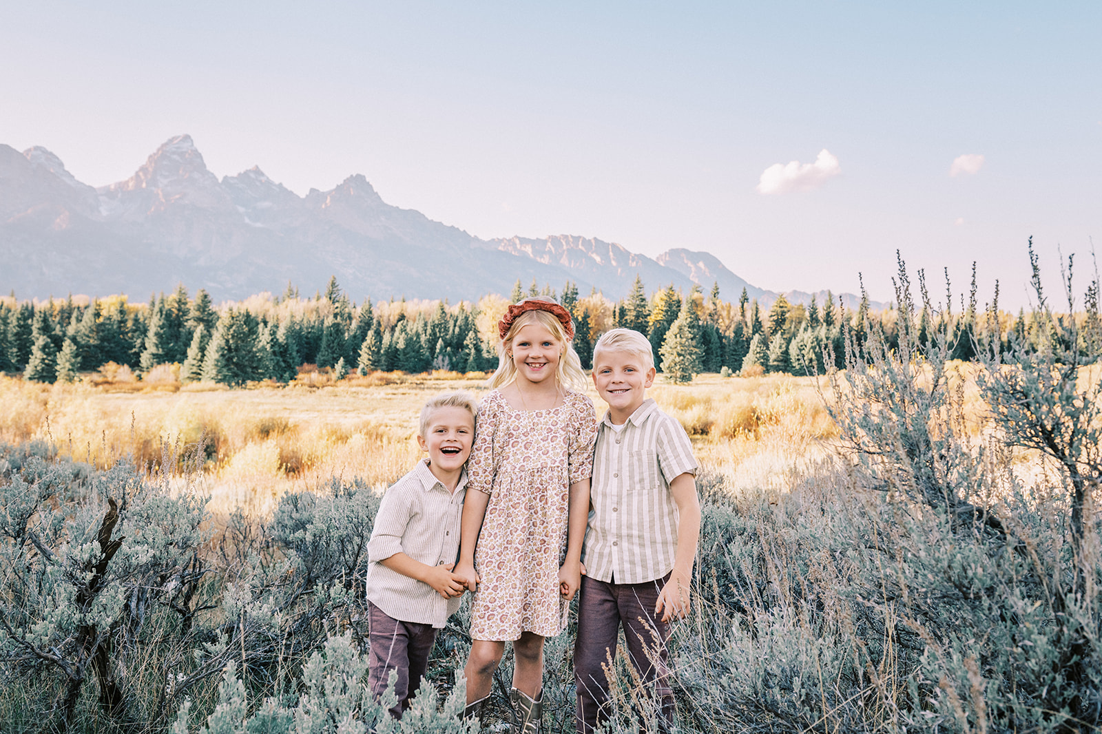Three giggling young siblings stand holding hands in a meadow of tall grass at sunset after finding things to do as a family in Jackson, WY