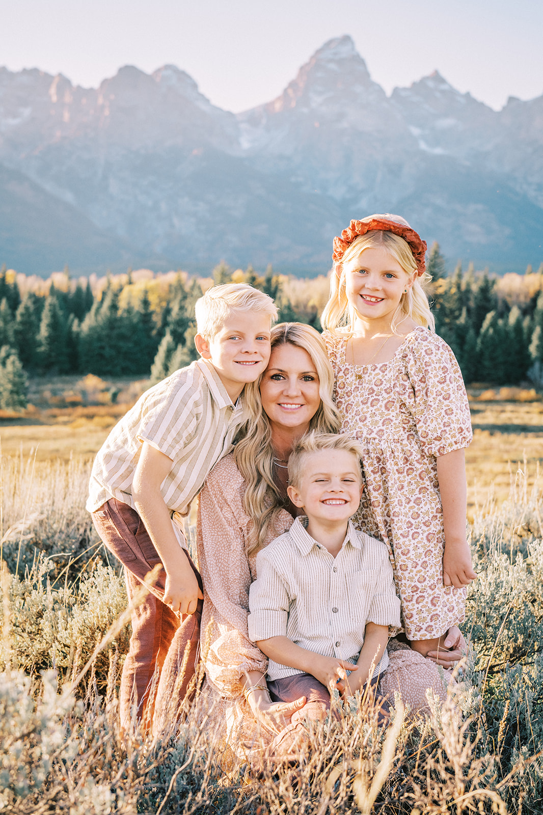 A happy mom kneels with her three young children hugging and sitting in her lap in a field after enjoying things to do as a family in Jackson, WY