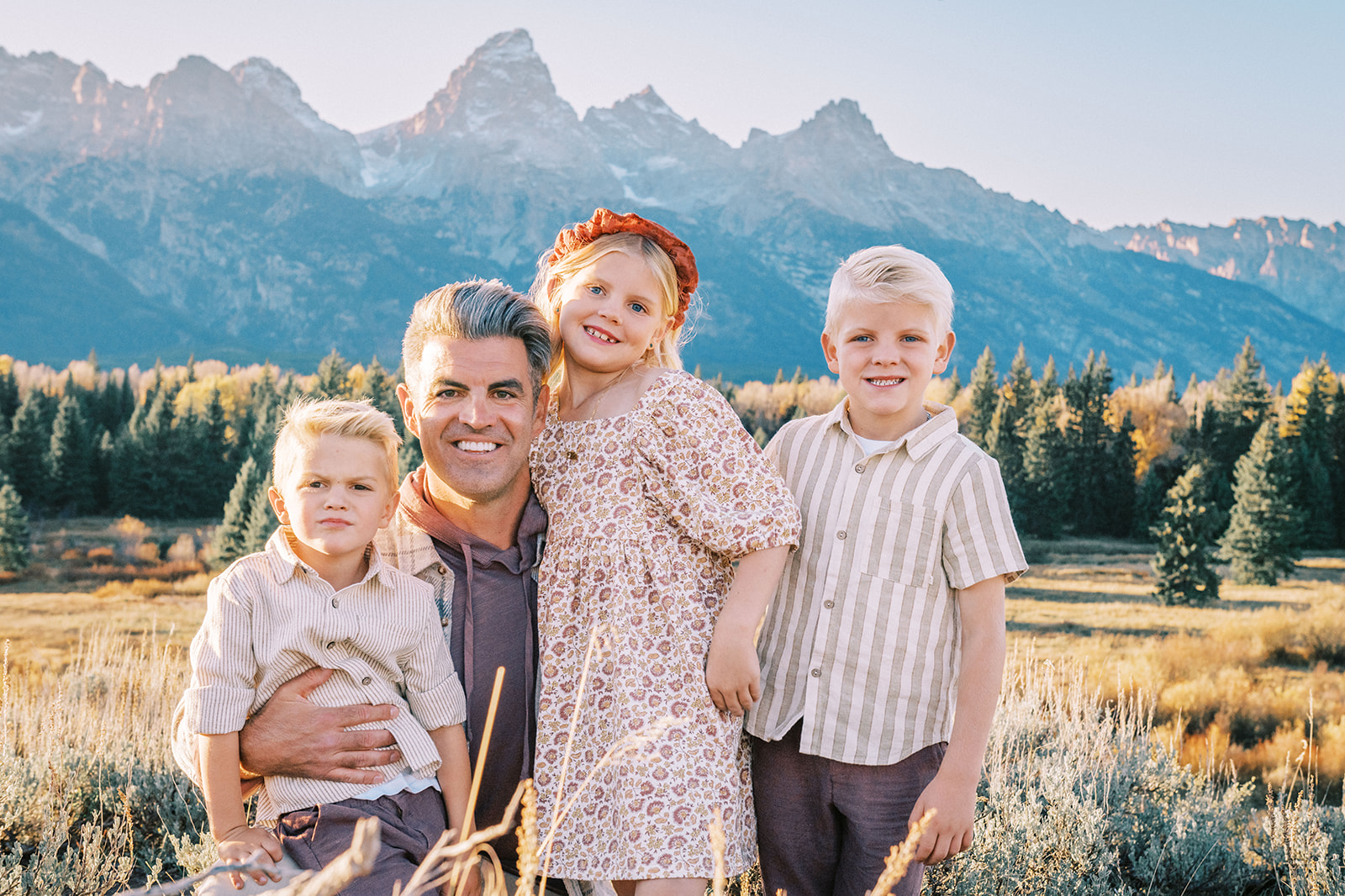 A happy dad kneels with his three young children in earth tones in a meadow after finding things to do as a family in Jackson, WY