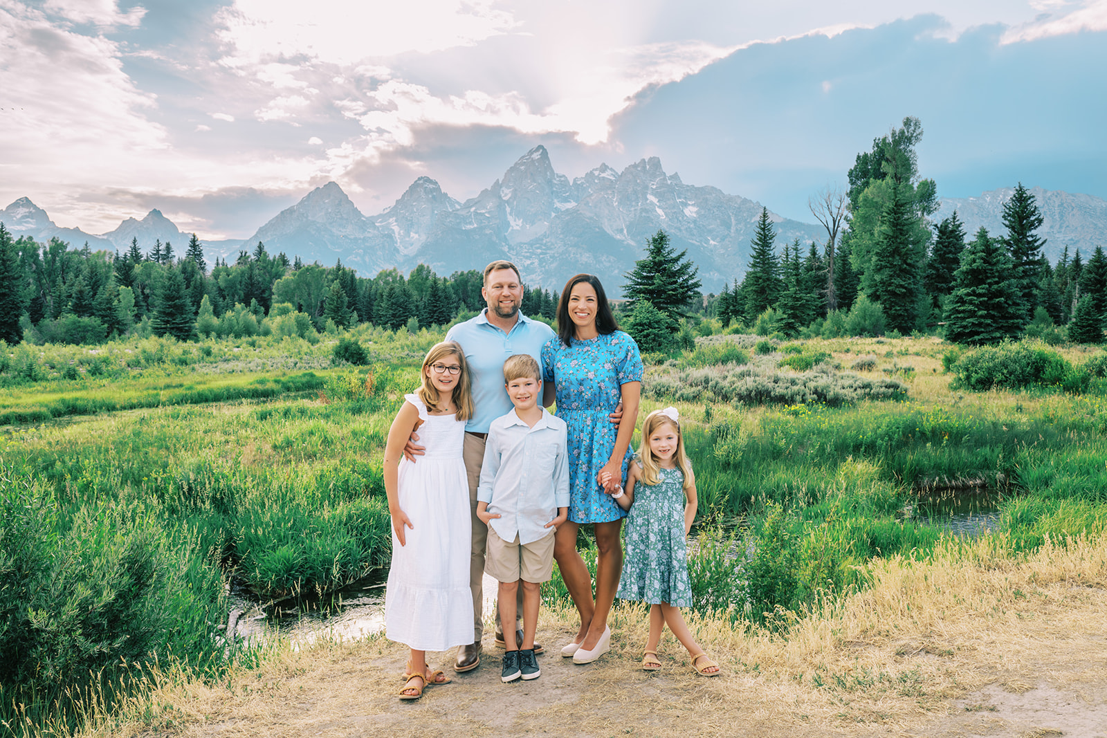 A mom and dad stand hugging and holding hands with their three young children in white and blue in a park at sunset by a river