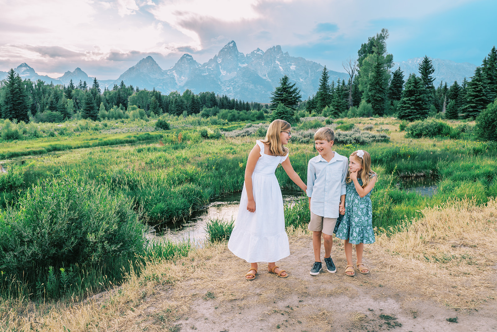 A young girl reaches to encourage her younger brother and sister to play in a trail by a river during things to do in spring in Grand Teton National Park with kids