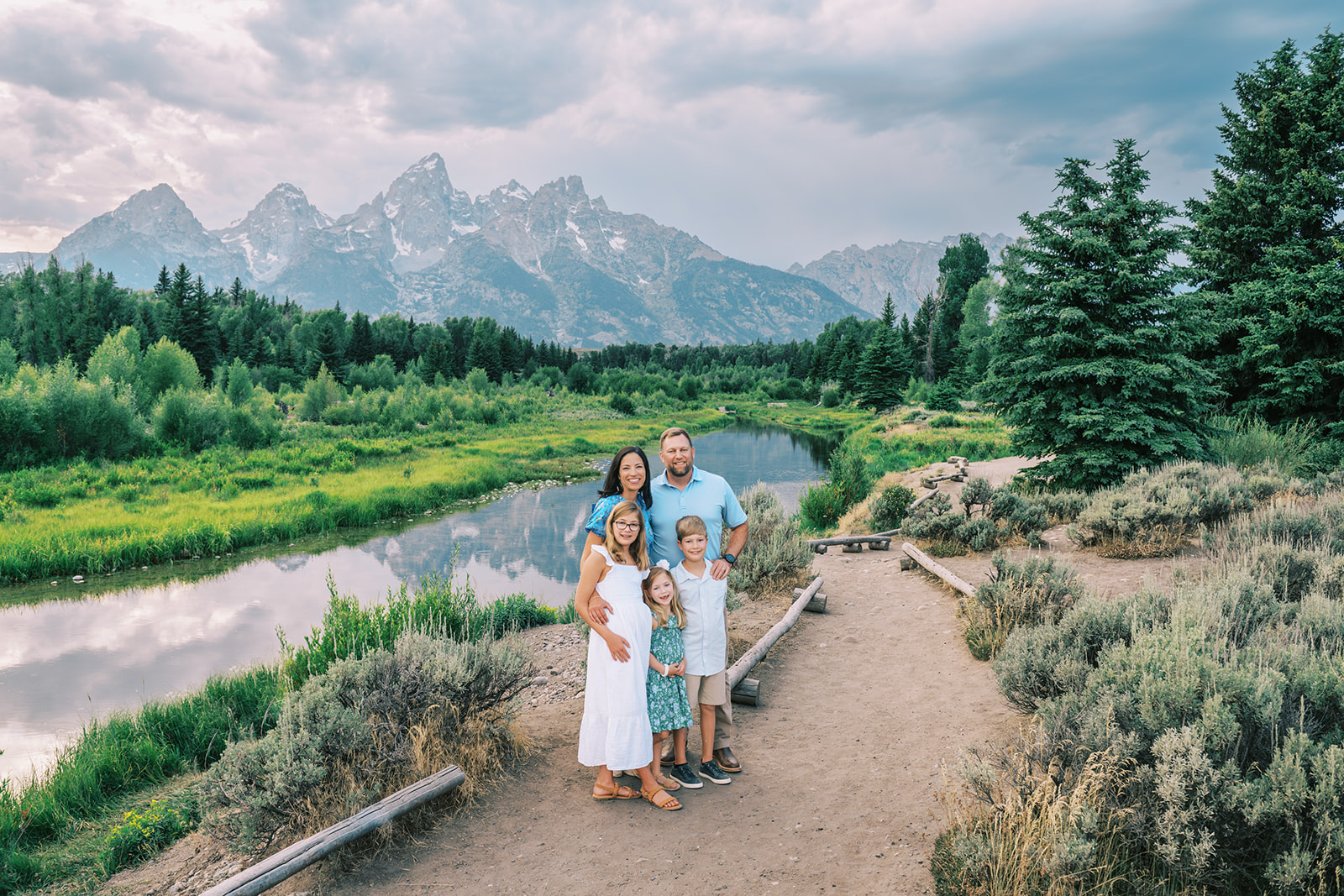 A happy mom and dad in blue stand with their three young children in white and blue in a park trail by a pond after finding things to do in spring in Grand Teton National Park with kids